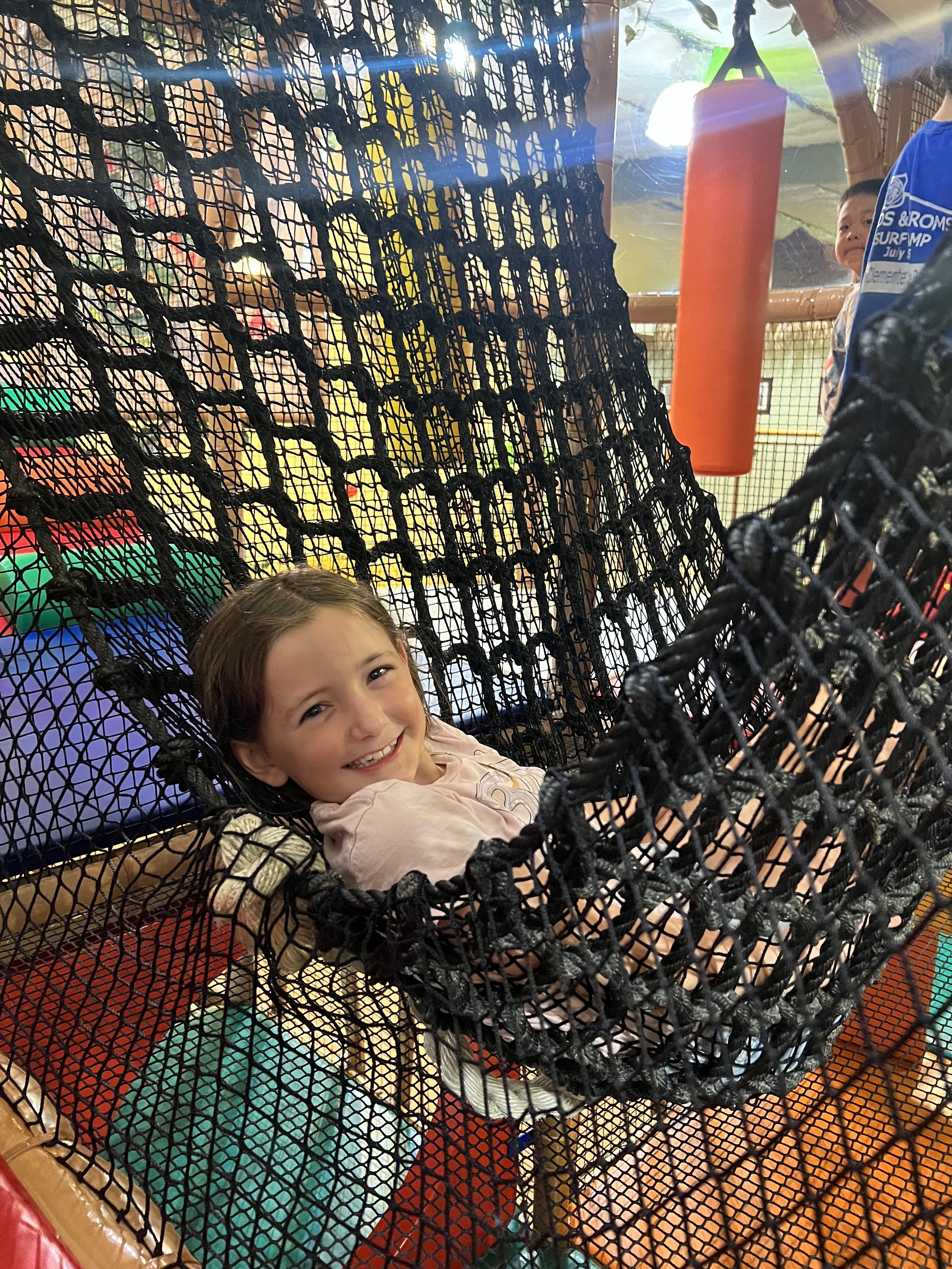 A smiling girl lounging in a black netted hammock inside the Safari Run play structure