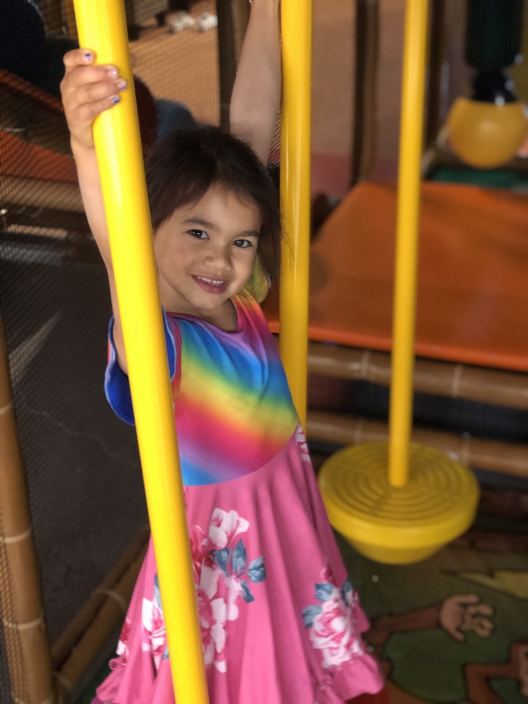 A girl in a rainbow dress hanging from a yellow bar at Safari Run