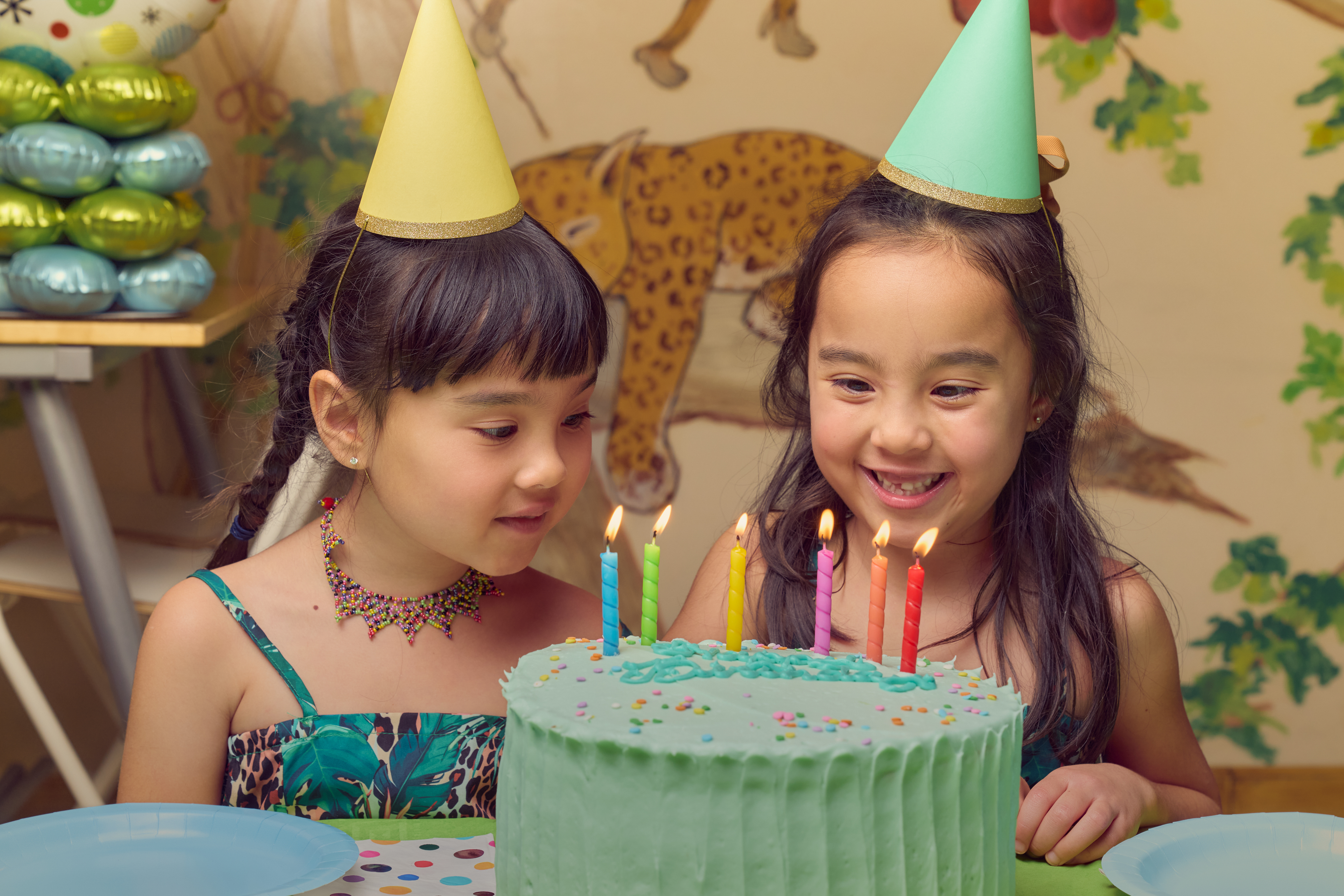 Two girls in party hats blowing out candles on a green birthday cake