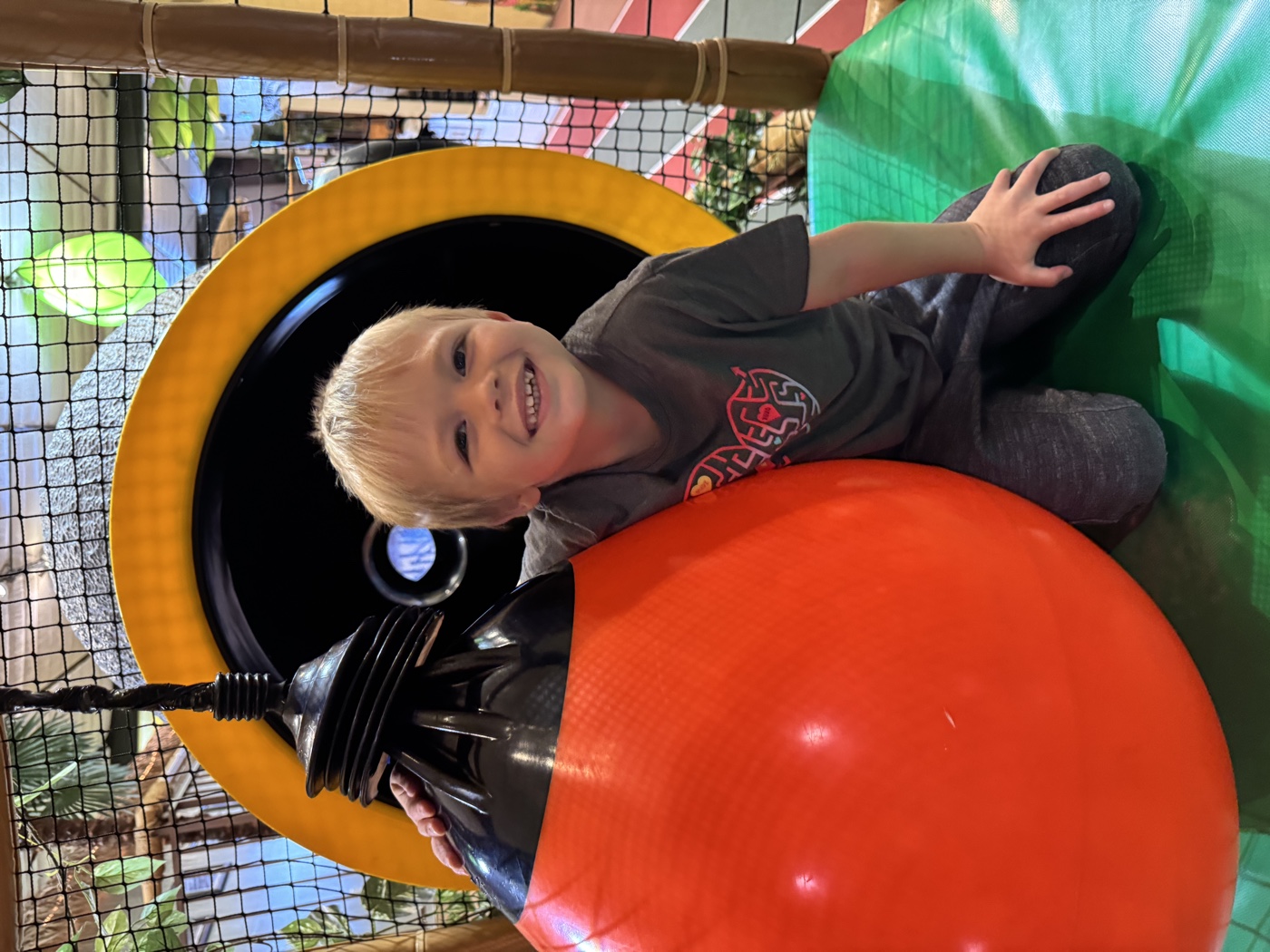 A smiling boy kneeling next to a large orange punching ball inside the Safari Run San Mateo play structure